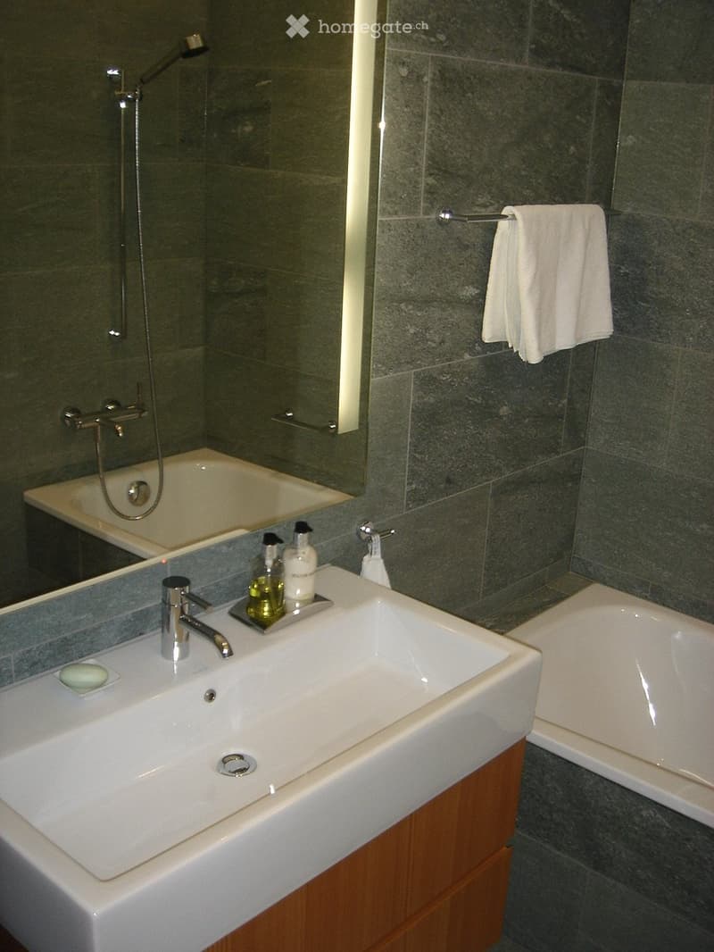 Modern bathroom with a white sink, chrome faucet, bathtub, and a towel hanging on a rack. Dark grey tiles line the walls and floor.