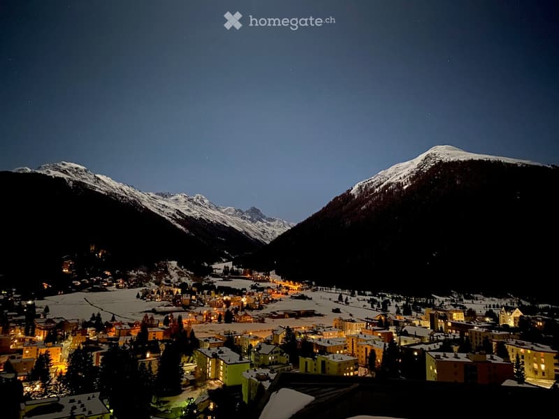 Nighttime view of a small village nestled in a snowy mountain valley, with illuminated buildings and surrounding snow-covered peaks.