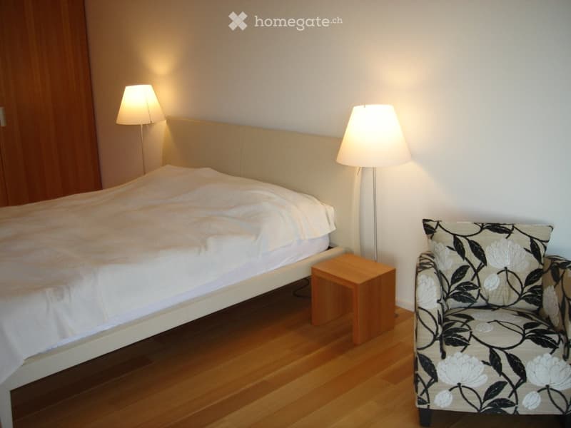 Cozy bedroom with a large bed, a patterned armchair, a small wooden stool, and two floor lamps.