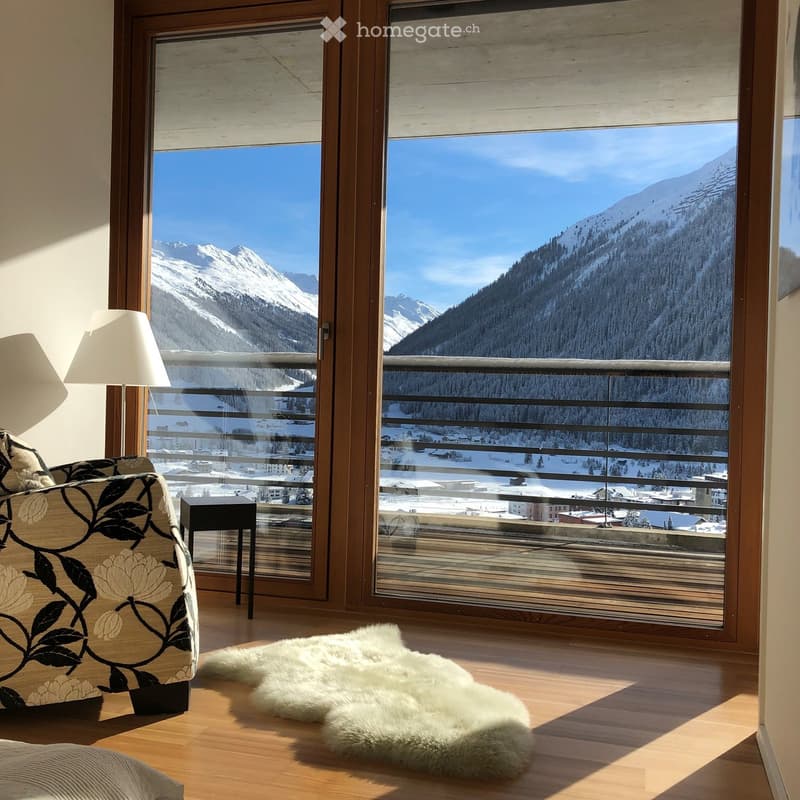 Bright room with large windows offering a scenic mountain view, featuring a patterned sofa, a small table, a floor lamp, and a white fur rug on the wooden floor.
