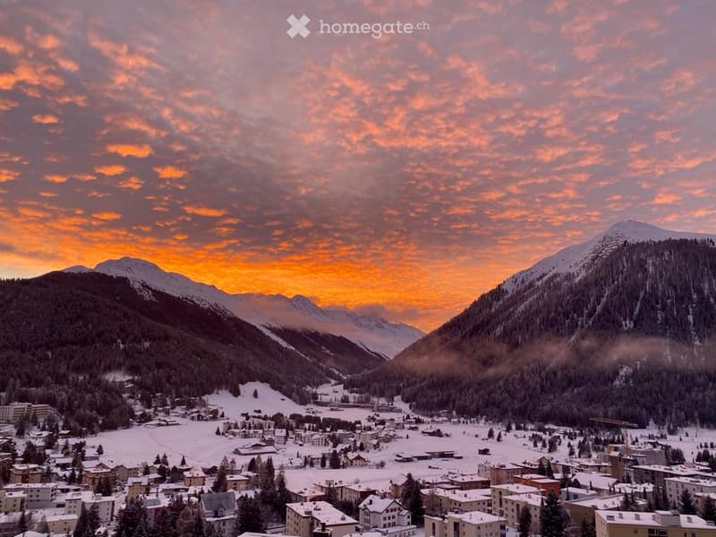 A panoramic view of a snow-covered town nestled in a valley surrounded by mountains, with a vibrant sunset sky.