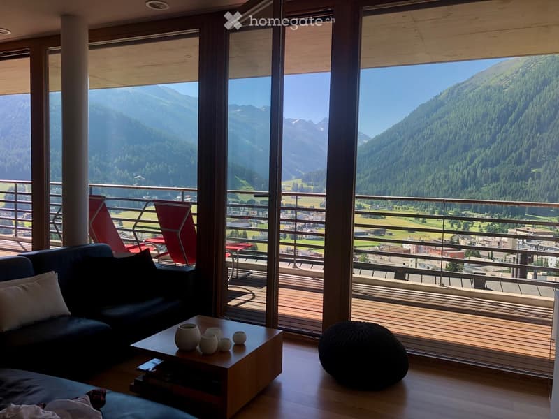 Modern living room with large windows offering a scenic mountain view, featuring a dark blue sofa, a wooden coffee table with decorative vases, and a round black ottoman.