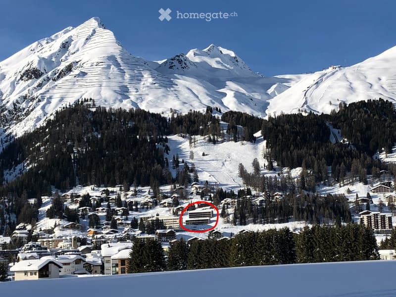 A snow-covered mountain range with a village nestled in the valley below, featuring multiple buildings and ski slopes.