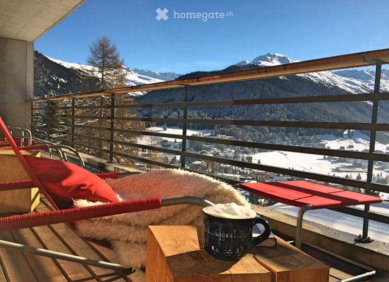 Balcony with a scenic mountain view, featuring a wooden table, a cup of hot chocolate, and a red lounge chair with a fur throw.