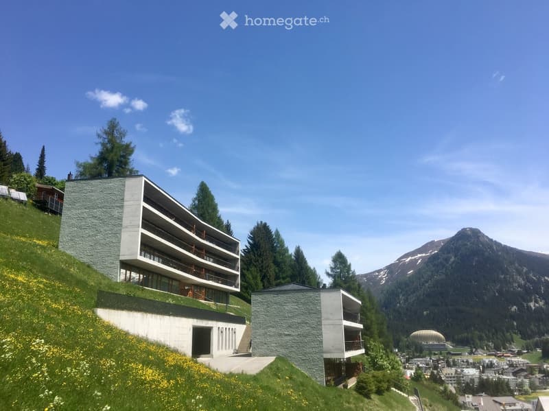 Modern multi-story building with balconies, situated on a hillside with a scenic mountain backdrop and surrounding greenery.