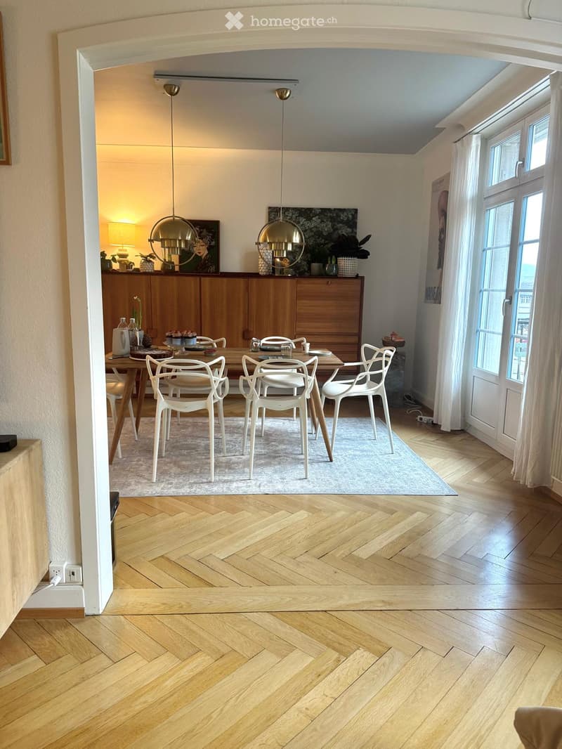 Dining area with a wooden dining table, white chairs, pendant lights, and a wooden cabinet with decorative items on top.