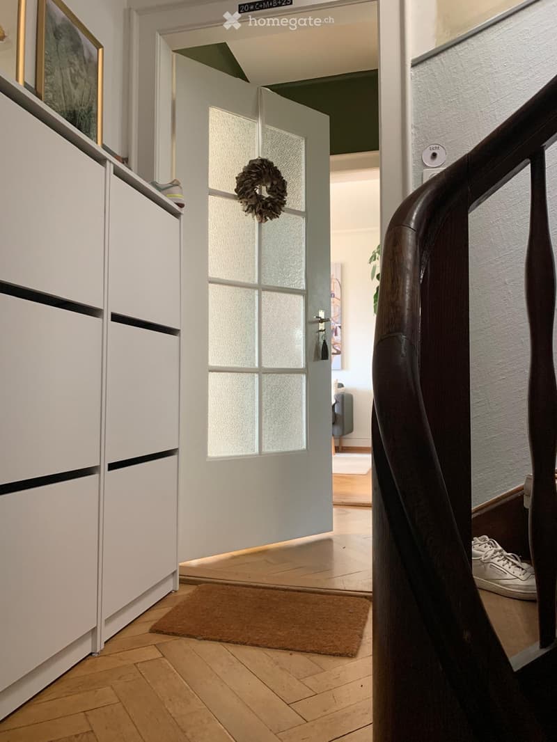Hallway with wooden flooring, white walls, a door with a wreath, and a small rug. A staircase is visible on the left, and a glimpse of a living area can be seen through the open door.