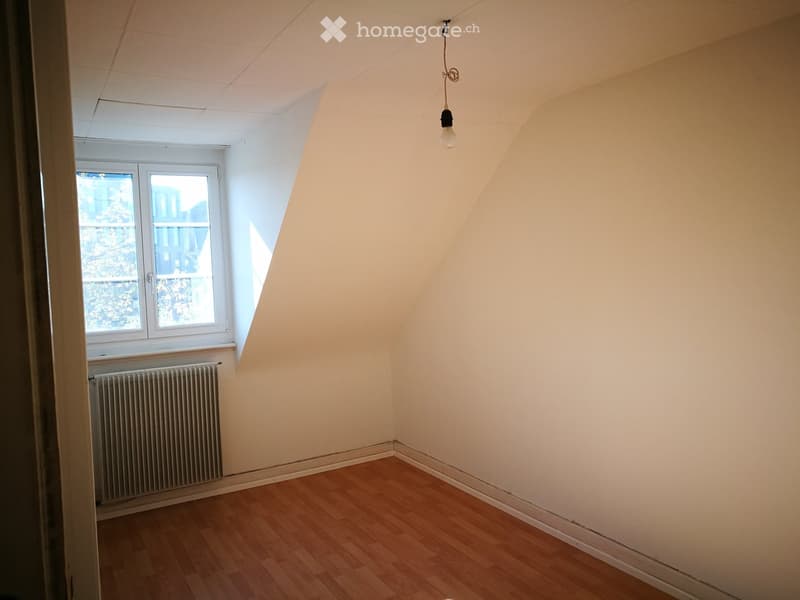 Empty room with wooden flooring, white walls, a single window with white frame, a radiator below the window, and a single hanging light fixture.