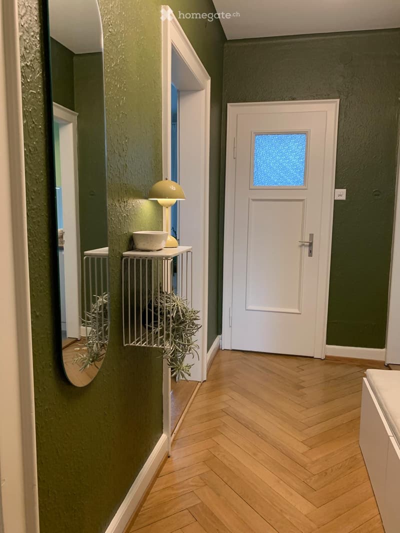 Hallway with green walls, herringbone wooden flooring, a small table with a lamp and plant, and a white door with frosted glass panel.