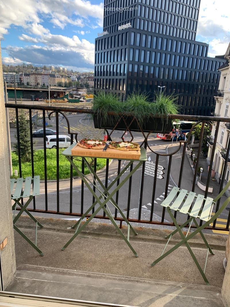 Balcony with a small table, chairs, pizza on a cutting board, and potted plants, overlooking a city street with traffic and buildings.