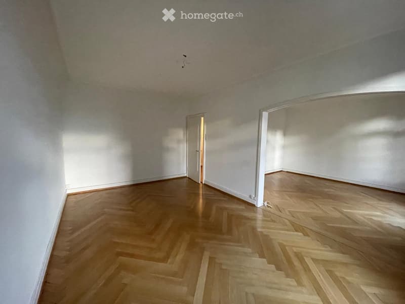 Empty room with wooden herringbone flooring and white walls, featuring a doorway leading to another space.