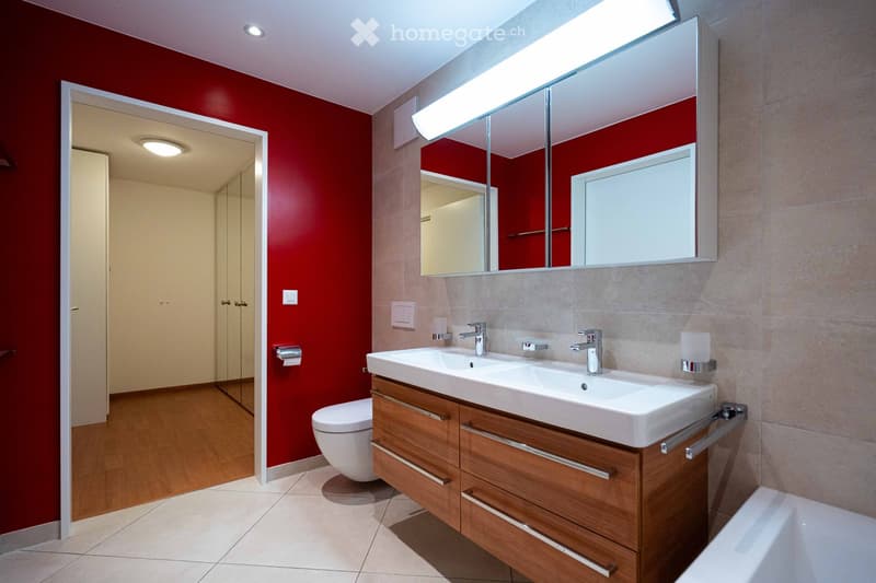 Modern bathroom with red walls, white sink, wooden cabinet, large mirror, and toilet visible.