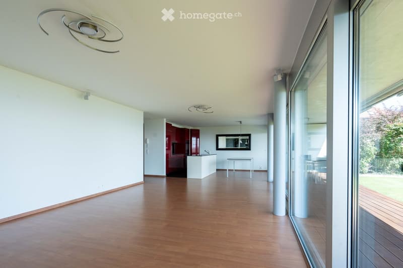 Modern, open-plan living and dining area with wooden flooring, large windows, and a contemporary kitchen area featuring red cabinets and a white island.