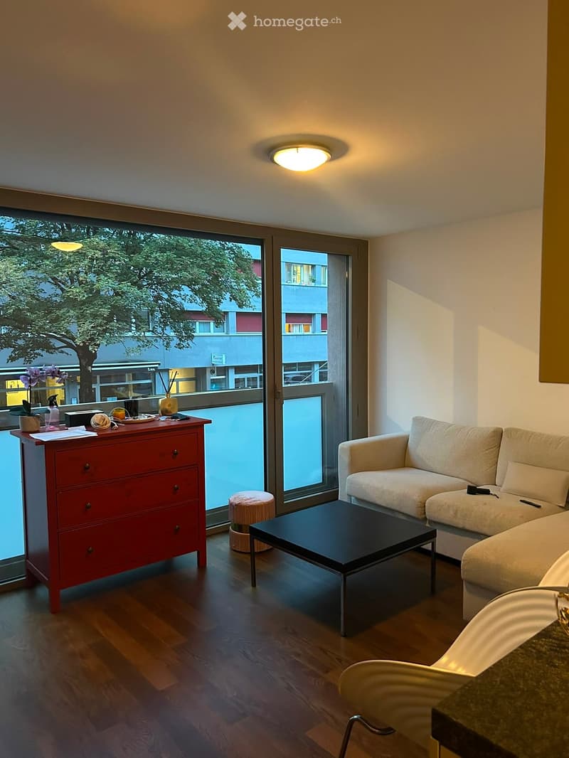Modern living room with a light-colored sofa, black coffee table, red chest of drawers, and a large window with a view of the street.