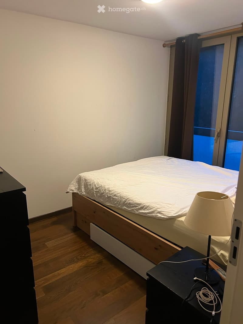 A small, modern bedroom with a wooden floor, a single bed with a white duvet, a nightstand with a lamp, and a window with dark curtains.