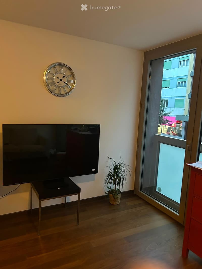 A modern living room with wooden flooring, a large wall-mounted television, a small table, a decorative clock, and a potted plant near the window.