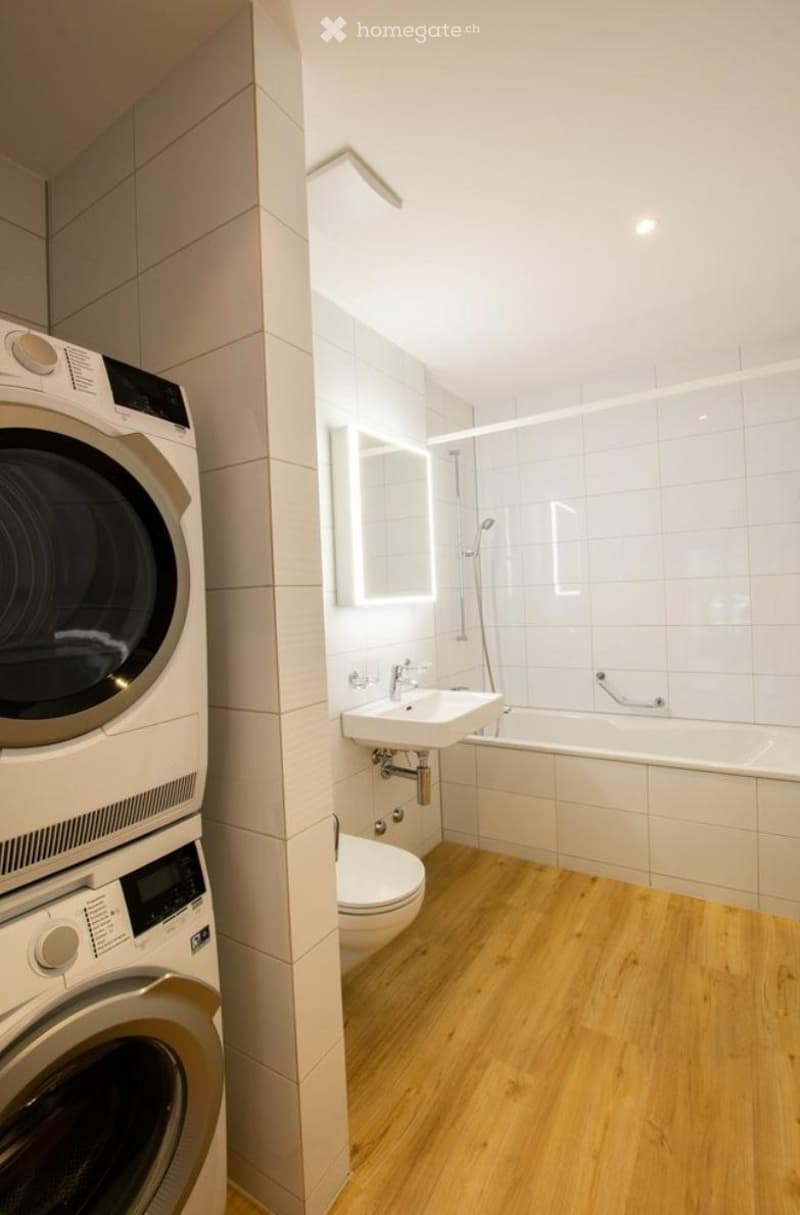 Modern bathroom with white tiles, wooden flooring, a sink, bathtub, toilet, and a built-in washing machine.