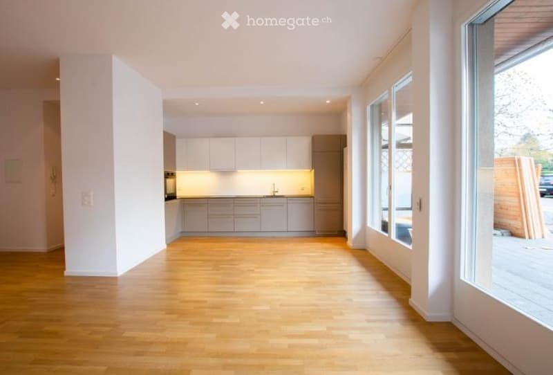 Open-plan space with a modern kitchen area featuring white cabinets and stainless steel appliances, connected to a large living area with wooden flooring and large windows.