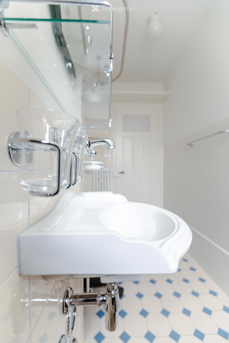 Modern bathroom with a white sink, chrome faucet, and a glass shelf above it. The floor has blue and white patterned tiles.