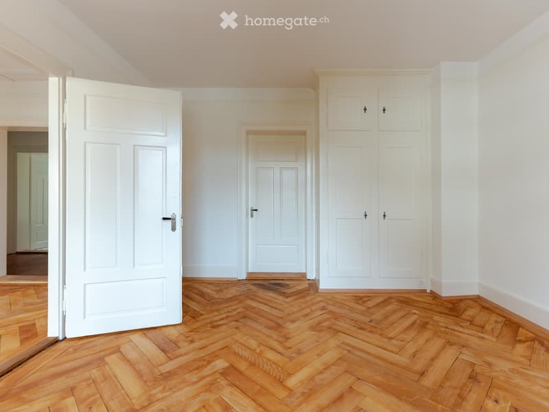 Empty room with wooden herringbone flooring, white walls, and several white doors.