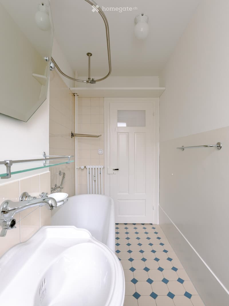 Bathroom with a white bathtub, sink, and modern faucet. Features a glass shelf, radiator, and a door with a frosted glass panel. The floor has a blue and beige tile pattern.