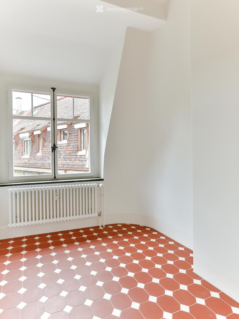 Empty room with white walls, hexagonal tile floor in shades of pink and red, large windows with view of rooftops, radiator below windows.
