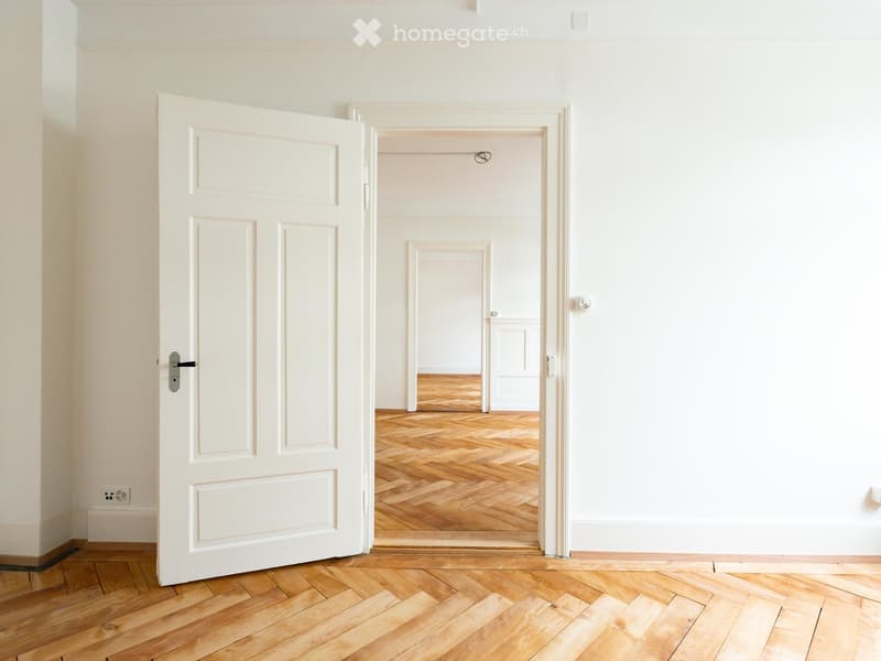 Empty hallway with white walls, wooden herringbone flooring, and a partially open door leading to another room.