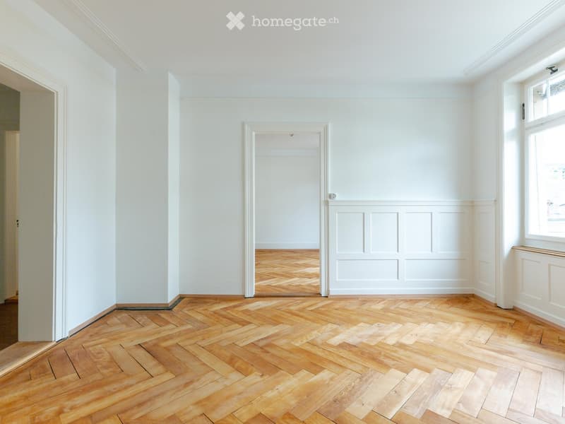 Empty room with light-colored walls, wooden herringbone floor, and a window with white frame. No furniture or equipment present.