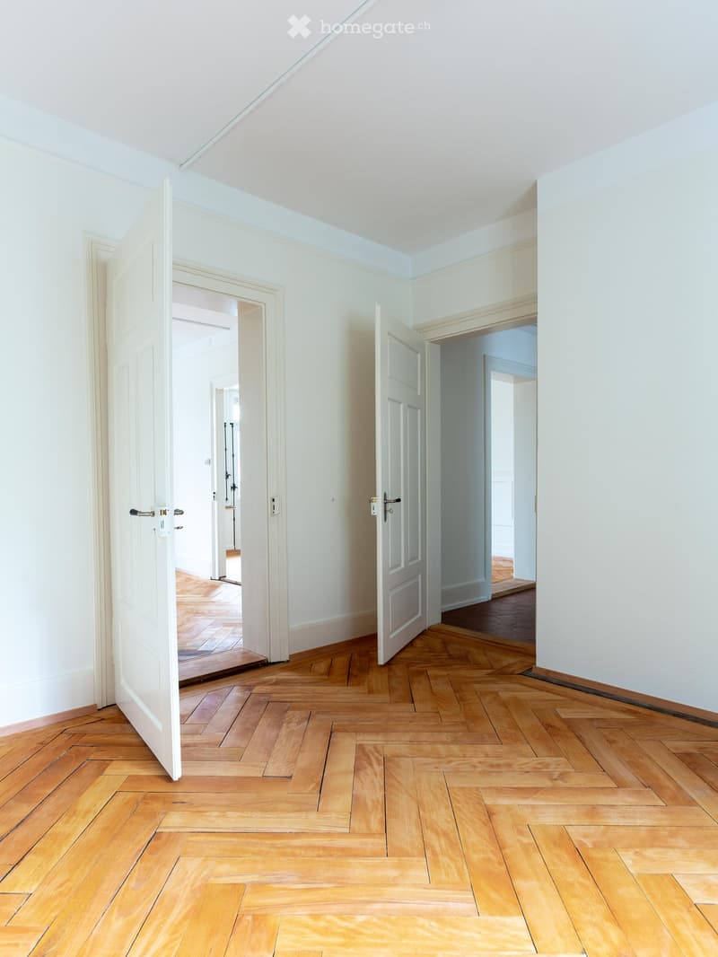 Empty hallway with wooden herringbone flooring, white walls, and open doors leading to other rooms.