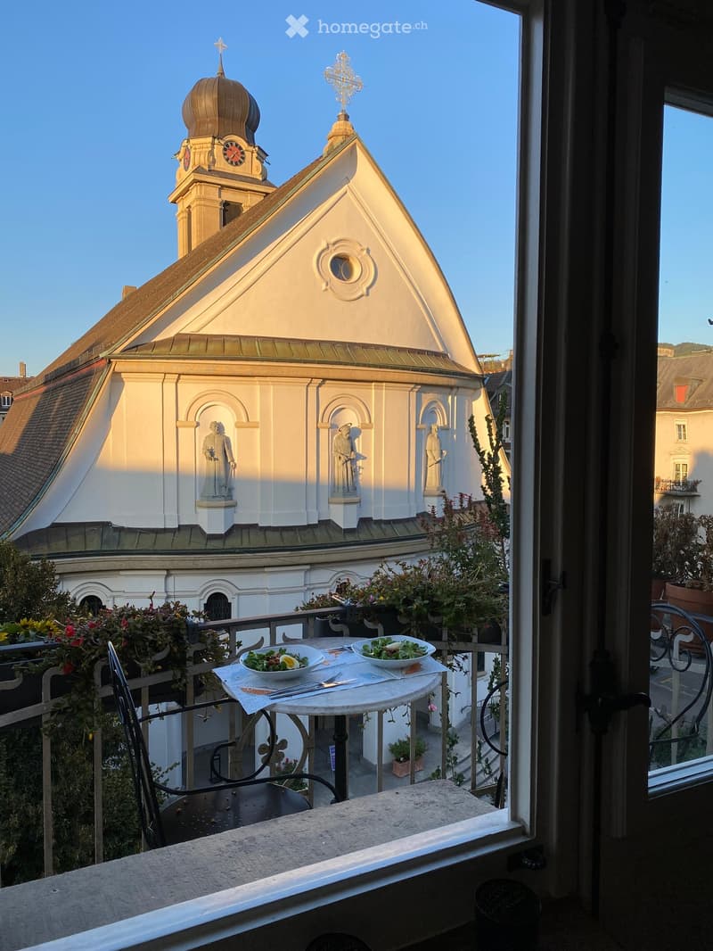 Balcony with a small round table, two chairs, and two bowls of salad, overlooking a historic church with a dome and statues.