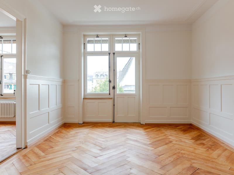 Empty room with light-colored walls, wooden herringbone flooring, and large windows with decorative grilles. Radiator visible on left wall.