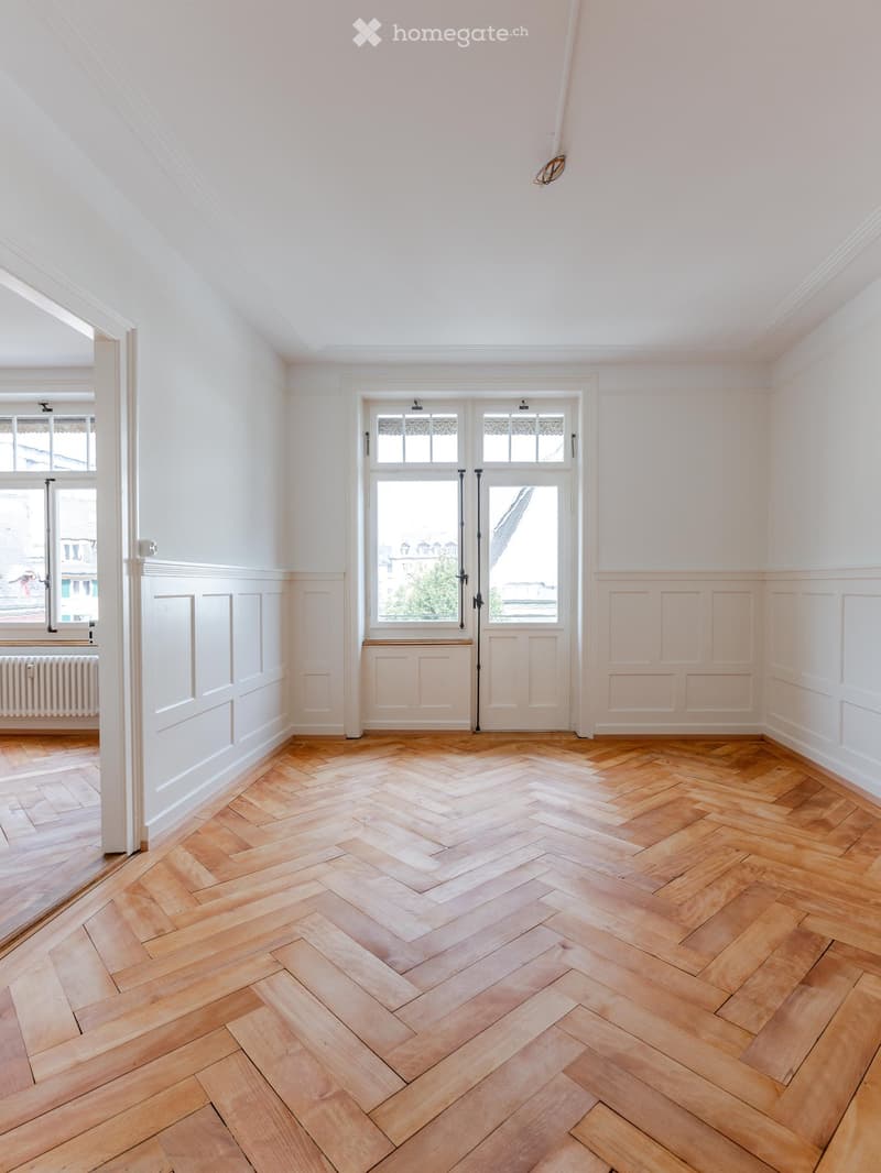 Empty room with light-colored walls, wooden herringbone flooring, and large windows with decorative glass panels.