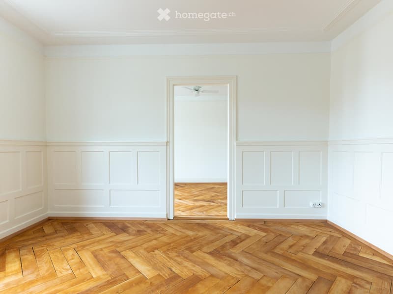 Empty room with light-colored walls, wooden herringbone flooring, and a white ceiling with a visible light fixture. The room features decorative paneling on the lower half of the walls and a doorway leading to another room.
