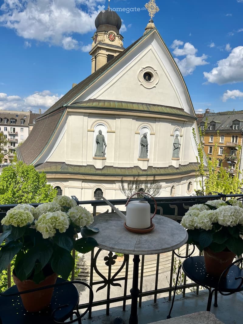 Balcony with a round table, two chairs, and potted flowers, overlooking a historic church with a dome and clock tower.