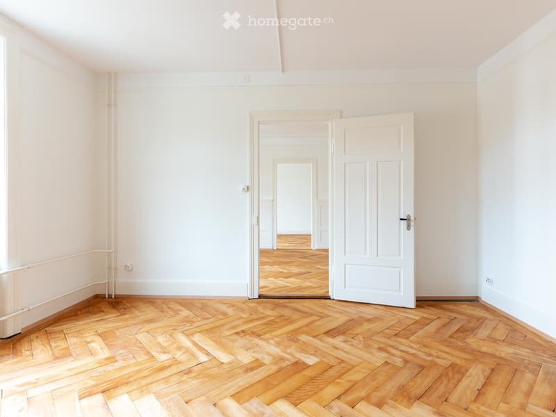 Empty room with light-colored walls and a herringbone wooden floor, featuring a white door leading to another room.