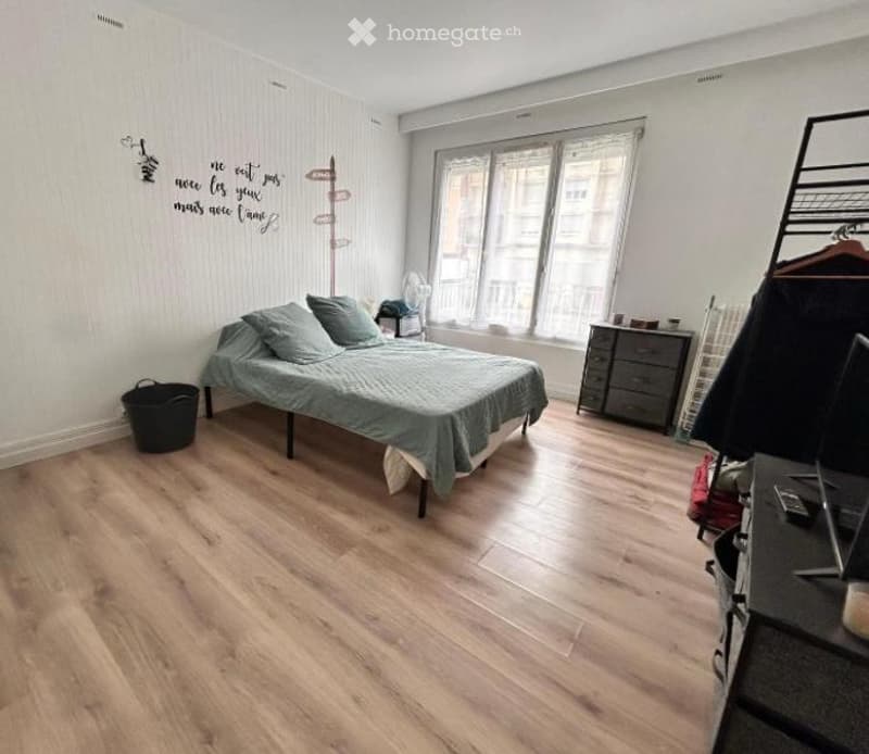 Bright bedroom with wooden flooring, a double bed with grey bedding, a nightstand, a fan, a chair, a dresser, a mirror, and a laptop on a stool.