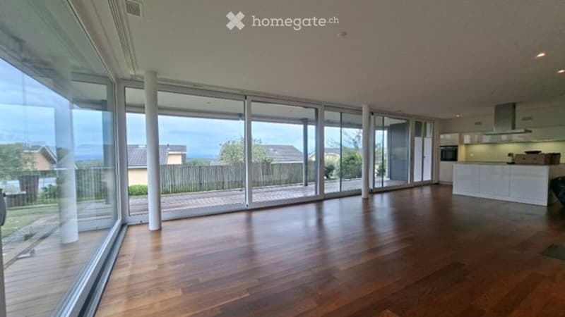Bright, open-plan space with large glass sliding doors leading to a patio, wooden flooring, and a modern kitchen area visible in the background.