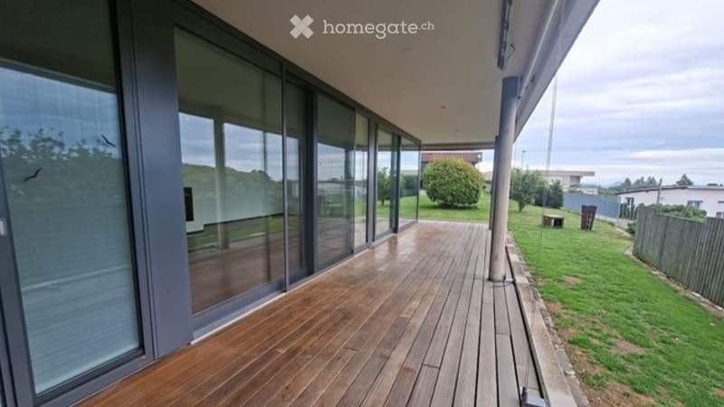 Modern wooden deck terrace with large glass sliding doors, surrounded by greenery and open space.