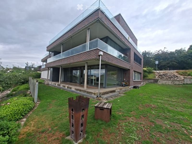 Modern two-story brick building with large glass windows and a raised ground floor, surrounded by a grassy area with a small wooden bench and a metal structure.
