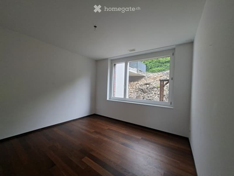 Empty room with wooden flooring and a large window showing a view of a rocky area and a raised ground floor outside.