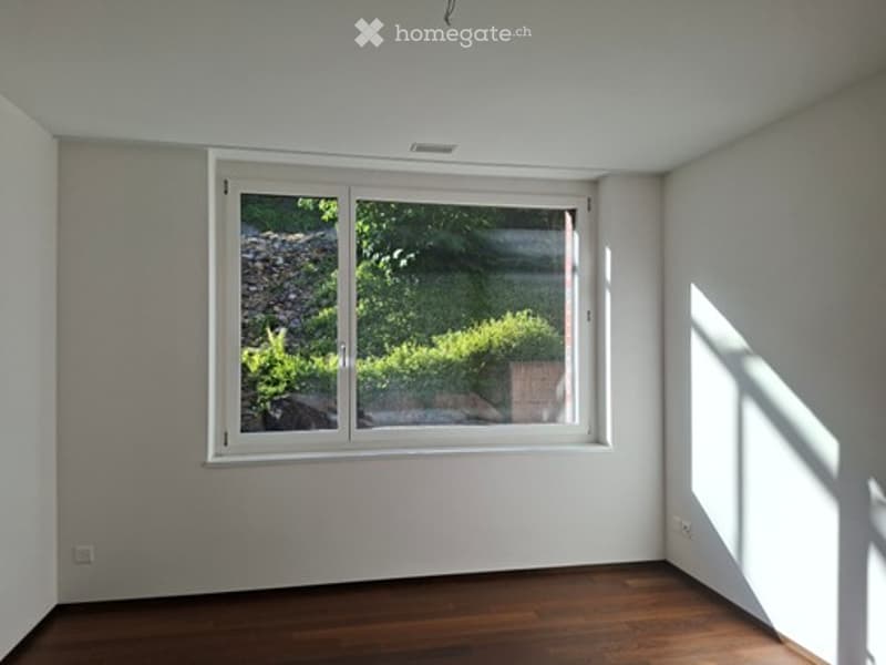 Empty room with wooden flooring, white walls, and a large window showing greenery outside.