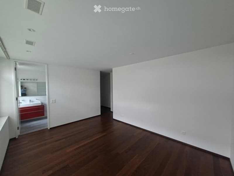 Empty room with wooden flooring and white walls, featuring a doorway to a red bathroom with a sink and mirror.