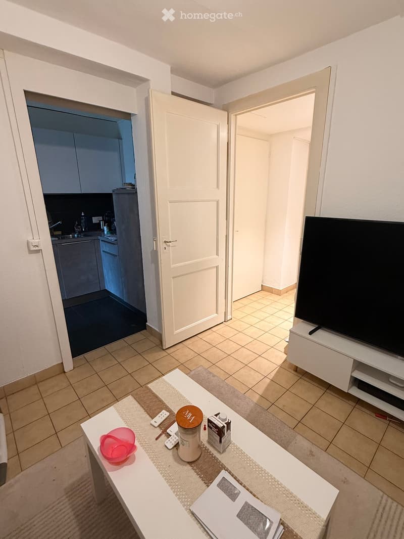 Living room with tiled floor, white coffee table with remote controls, cup, bowl, and TV stand with flat-screen TV. Open door leads to kitchen area with cabinets and sink.