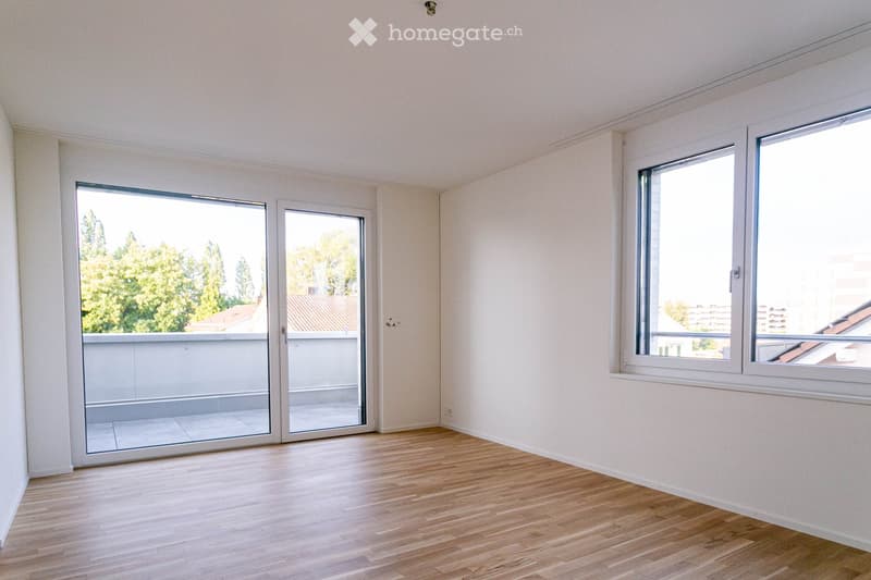 Empty room with light wooden flooring, white walls, large windows, and sliding glass doors leading to a balcony.