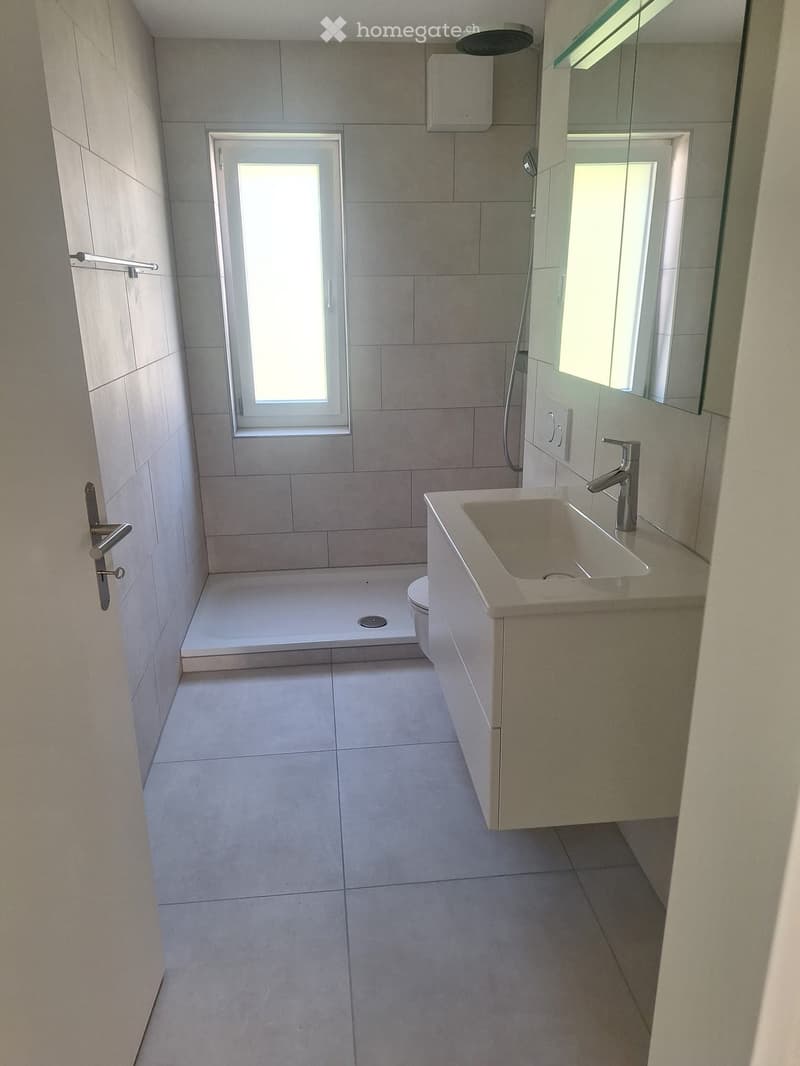 Modern bathroom with a large sink, mirror, shower, and toilet. The space features tiled walls and floor, a window for natural light, and a sleek design.