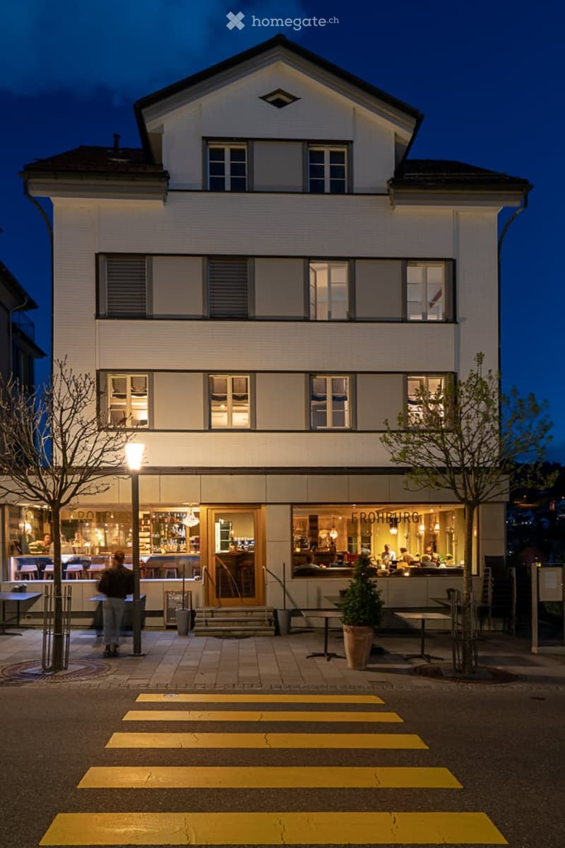 Three-story building with illuminated windows and a modern facade, featuring a restaurant or bar area with outdoor seating and a pedestrian crossing in front.