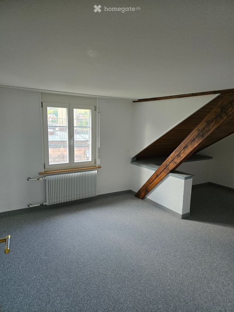 Empty room with grey carpet, white walls, a window with a radiator below, and a wooden staircase leading upwards.
