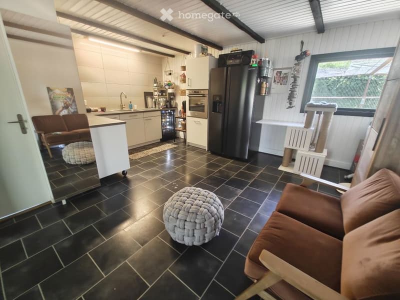 Open-plan living and kitchen area with dark tiled flooring, white cabinets, a sofa, a round stool, a cat tree, and a kitchen island with bar stools.