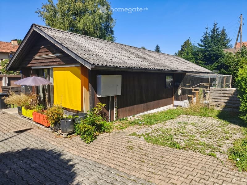 A small, single-story house with a dark wooden exterior and a grey tiled roof. The front features a yellow accent wall, a small patio with potted plants, and a gravel driveway. A wooden fence encloses a small garden area with additional plants and outdoor