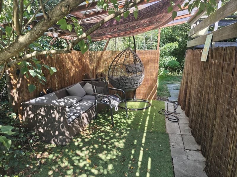 A cozy outdoor patio with a wicker sofa, a hanging egg chair, a small table, and a chair, surrounded by bamboo fencing and artificial grass.